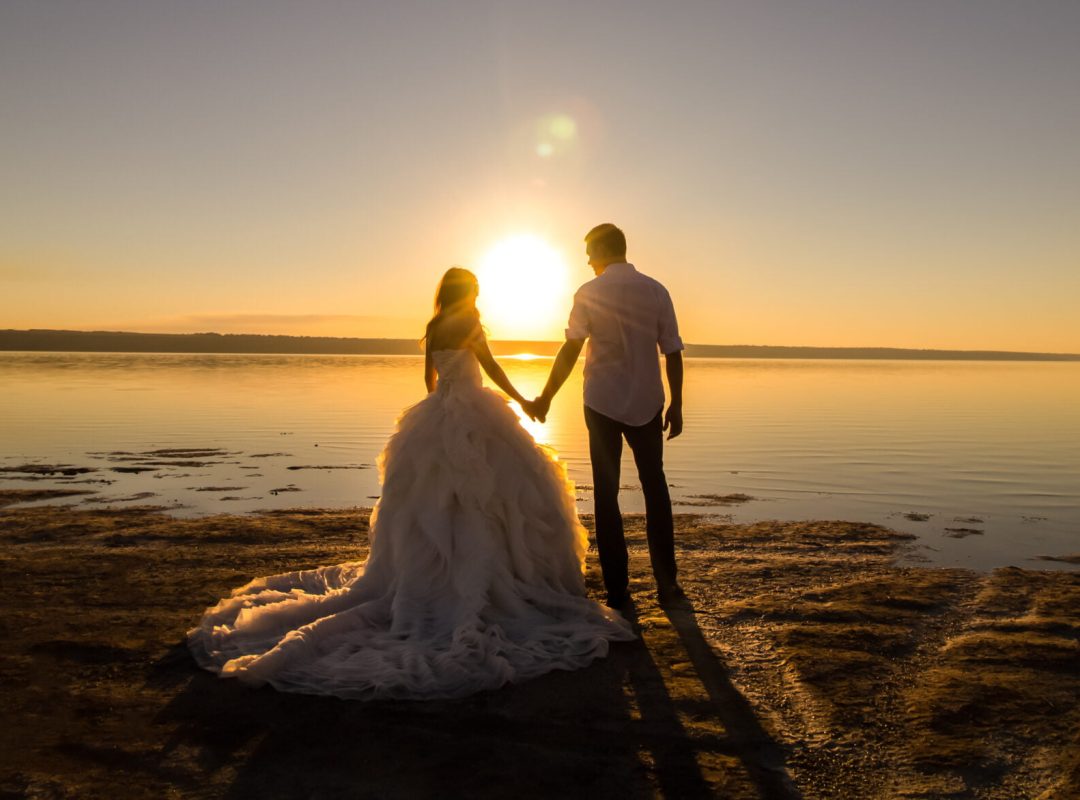 Wedding couple on the sea beach watching sunset. Sunny summer photo. Bride with hair down in off shoulder dress with train. Ocean romantic ceremony. Seaside sunrise love story. Honeymoon.